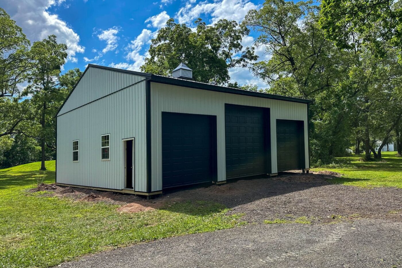 Black and white garage with three garage doors and two windows and a door on the side facing the camera set in a gravel driveway with green grass and blue sky
